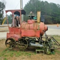 Old, rusted steam locomotive with visible wear, displayed in a grassy roadside area. A person sits smiling inside the cab, looking through the window. Surrounding elements include a traffic cone, trees, and a nearby road, giving it the feel of a quirky roadside display or decorative relic.