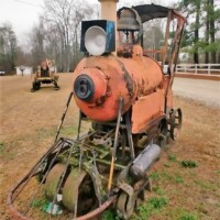 Rusty, weathered steam engine-like sculpture with a cylindrical orange body, large front headlamp, and exposed pipes, resting on wheels along a dirt path. Surrounded by trees and a second aged machine in the distance.