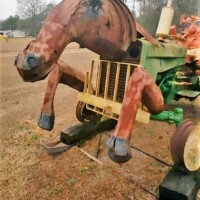 When the horsepower isn’t just metaphorical—it’s got hooves and hydraulics Side view of a whimsical sculpture blending the front half of a painted horse with the rear half of a green tractor, complete with large yellow wheels and visible engine parts. The sculpture stands in a grassy clearing, with trees and shrubs in the background.
