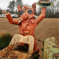 Metal sculpture resembling a pig-like creature with glasses, constructed from repurposed parts and perched atop a rusty tractor. The figure holds a hat in one hand, with trees and a house visible in the background.