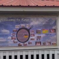 Large welcome sign for Rose Hill, North Carolina, displayed under a red-roofed shelter, featuring a graphic of the World’s Largest Frying Pan and local attractions.