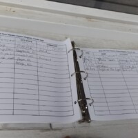 Open visitor logbook for the World's Largest Frying Pan, displayed on a white surface with a blue pen resting beside a three-ring binder filled with handwritten guest entries.