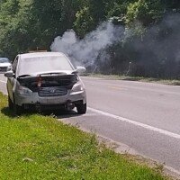 A white car pulled over on a roadside with smoke rising from the engine area, trees behind