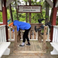 Person in a beanie and hoodie braces face-first inside the Angus Barn Kicking Booth, feet comically airborne behind as the legendary leather boots prepare to strike.