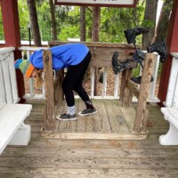 Person in a blue hoodie leans into the Kicking Booth at Angus Barn, perfectly positioned for comedic impact as the swinging boots await behind under a garden pavilion.