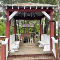 Wooden gazebo with white benches and red trim at the Angus Barn, flanked by lanterns and trees, offering a quiet resting spot beside the chaos of the Kicking Booth.