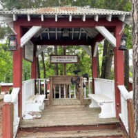 Wooden gazebo with white benches and red trim at the Angus Barn, flanked by lanterns and trees, offering a quiet resting spot beside the chaos of the Kicking Booth.