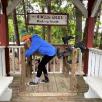 Person in a blue hoodie and rainbow hat stands solemnly in front of the Angus Barn Kicking Booth, fully committed to a self-inflicted boot-to-butt moment under a wooden arbor with benches nearby.