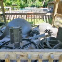 Outdoor display of historic sugar mill machinery, including gears and iron components arranged on gravel within a wooden enclosure, surrounded by trees and a grassy clearing at DeLeon Springs