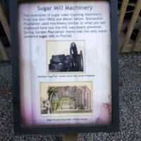 Informational sign titled 'Sugar Mill Machinery' at DeLeon Springs, describing 1800s steam-powered sugar processing equipment donated by the Louisiana State Penitentiary, with historic illustration and photo of the machinery.