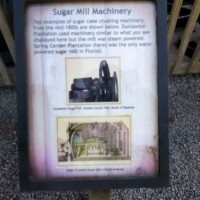 Informational sign titled 'Sugar Mill Machinery' at DeLeon Springs, describing 1800s steam-powered sugar processing equipment donated by the Louisiana State Penitentiary, with historic illustration and photo of the machinery.