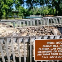 Fenced-off ruins of an 1850s–1870s sugar mill at DeLeon Springs with a sign reading: 'Sugar Mill Ruins. Florida law prohibits disturbing or removing anything from this site.