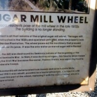 Interpretive sign titled 'Sugar Mill Wheel' explaining the history of DeLeon Springs' iron water wheel used to grind sugar cane in the 1800s, including its destruction in a 1901 hurricane and survival as the last remnant of the original sugar mill.