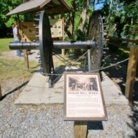 Large historical sugar mill gear system on display at DeLeon Springs, featuring two iron gears mounted on wooden beams, with an informational sign in front and a sheltering wooden pavilion in the background, surrounded by trees.