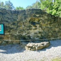 Section of historic stone wall remains at DeLeon Springs, partially roped off and accompanied by an informational sign describing the site's sugar mill foundations, set in a grassy clearing under trees.