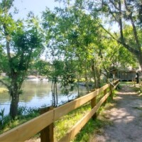 Tranquil path beside a reflective spring at DeLeon Springs, bordered by a wooden fence on one side and greenery on the other, with a bench and rustic cabin in the background under tall trees.