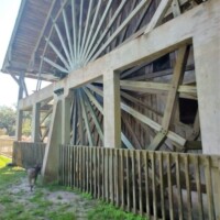 Large vertical wooden water wheel at DeLeon Springs, attached to a rustic wooden mill structure with support beams and a surrounding fence, set against a grassy, sunlit backdrop.