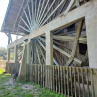 Large vertical wooden water wheel at DeLeon Springs, attached to a rustic wooden mill structure with support beams and a surrounding fence, set against a grassy, sunlit backdrop.