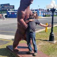 Zach posing with a giant bear statue outside a Discount Fireworks store, standing on a red platform with one arm around the bear and the other extended, blending roadside kitsch with travel charm.