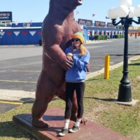 Nancy posing with a giant brown bear statue outside a roadside attraction, wearing a yellow beanie and blue sweatshirt, with colorful flags and a parking lot in the background.