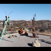 An outdoor art installation featuring a large green praying mantis sculpture, a rusted metal tree-like structure, and two spherical objects, set against a hilly landscape with scattered houses.
