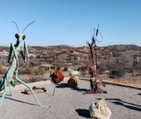 An outdoor art installation featuring a large green praying mantis sculpture, a rusted metal tree-like structure, and two spherical objects, set against a hilly landscape with scattered houses.