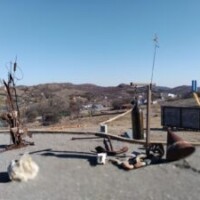 An outdoor display featuring rusted metal structures, pipes, and other weathered objects arranged on a flat surface. In the background, there are rolling hills, scattered buildings, and a clear blue sky. A small sign is visible, but its text is unreadable.