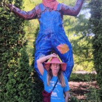 Nancy posing with a giant scarecrow in blue overalls and a straw hat, surrounded by tall green bushes and garden greenery.