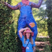 Nancy posing with a giant scarecrow in blue overalls and a straw hat, surrounded by tall green bushes and garden greenery.