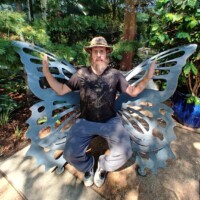 Zach sitting on a metallic butterfly wing bench in a garden, wearing a black graphic tee and white sneakers, blending nature with roadside whimsy.