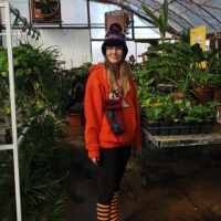 Nancy standing in a greenhouse wearing an orange hoodie and striped socks, surrounded by lush potted plants and natural light.