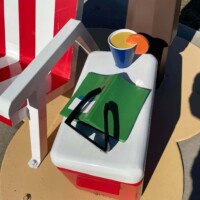 Close-up of a red-and-white cooler resting on a circular platform, topped with a green folder, black sunglasses, and a vibrant orange-yellow drink in a cup. A striped red-and-white chair and faux palm tree pole frame the scene, channeling a laid-back tropical vibe in a parking lot setting.