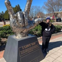 Towering metallic sculpture of a hand making the “I love you” gesture in American Sign Language. Mounted on a pedestal with a plaque reading “I LOVE YOU,” the piece celebrates inclusivity and connection. A person in a black outfit and beanie mirrors the sign beside it, adding a human echo. Set against a backdrop of trees and a bright blue sky along a paved walkway, the scene feels heartfelt and iconic
