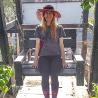 Nancy sitting on a wooden swing in a garden setting, wearing a wide-brimmed hat and colorful socks, surrounded by greenery and rustic charm.