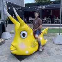 Zach sitting on a giant yellow horned fish sculpture outside a restaurant with string lights and outdoor diners, blending whimsy with roadside charm.