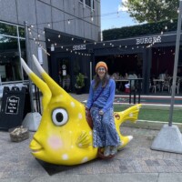 Nancy sitting on a giant yellow horned cowfish sculpture outside CowFish Sushi Burger, wearing a blue top and orange beanie, with string lights and diners in the background