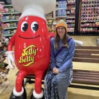 Nancy sitting beside the Jelly Belly mascot inside a candy store, surrounded by shelves of colorful jelly beans and sweet chaos.