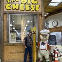 Zach standing beside a giant chef mouse statue at The Big Cheese, under a height-check sign, with kielbasa and cheese curd signs in the background.