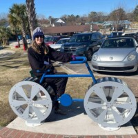 Large blue metal bicycle sculpture with exaggerated white-spoked wheels, designed as an interactive art piece. A person wearing a winter hat and sunglasses sits on the bike, adding a playful human touch. Behind them, palm trees, parked cars, and open sky hint at a sunny park setting, creating a whimsical contrast between seasons and scenery.