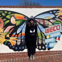 Vibrant mural painted on a brick wall in Columbia, SC, featuring a large butterfly with multicolored wings. Each wing includes detailed illustrations of flowers, a flamingo, a pelican, and a building labeled “WECO.” The mural also displays the artist’s tag “GMP Murals” and Instagram handle “@goodmurals4you.” A person stands centered in front of the mural, positioned so the butterfly wings appear to extend from their back, creating a playful and immersive effect.