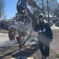 Outdoor photo featuring a person in a black sweatshirt, beanie, and black pants standing beside a whimsical metal sculpture shaped like a seahorse. The sculpture is constructed from repurposed metal parts, evoking a steampunk-meets-nautical vibe. Behind them, a mix of greenery, parked cars, and a public sign with the letter “H” suggests a relaxed, urban setting. The interaction between human and art creates a quirky yet thoughtful contrast—like spotting magic in a scrapyard.