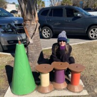 Outdoor installation featuring three oversized industrial spools wrapped in vibrant yellow, purple, and orange materials. A person sits nearby under a tree in a sunlit parking lot, adding a quiet human element. A green cone-shaped object stands beside the spools, enhancing the surreal, playful feel. Parked cars and leafy trees frame the scene.