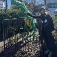 Outdoor scene featuring a person dressed in black standing next to a green T-Rex sculpture enclosed within a black metal fence. This T-Rex has yellow accents and a slightly different stance from the previous one. The person rests one arm casually on the fence, the other by their side, framed by mulch-covered ground, leafy trees, and a Sysco truck in the background. The vibe is roadside curiosity meets prehistoric photobomb, with a splash of ironic cool.