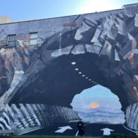 Large-scale mural on the side of a building in Columbia, South Carolina, depicting a rocky tunnel carved through a mountain with a road leading inward. Realistic painted details include jagged stone textures, ceiling lights along the tunnel interior, and directional white arrows on the road. In the distance, a scenic landscape with layered mountains and a warm sunset creates an optical illusion of depth.