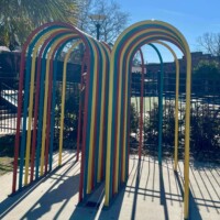 Colorful outdoor sculpture in Columbia, SC featuring a series of metal arches painted red, yellow, blue, and green, arranged to form a tunnel-like walkway. The vibrant structure casts playful shadows on the ground and is surrounded by a fence and greenery, creating a dynamic installation space.