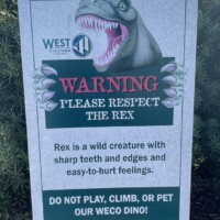 Playfully stern warning sign from “West Hills” featuring a cartoon-style green T-Rex with yellow accents. The sign reads: “WARNING — PLEASE RESPECT THE REX. Rex is a wild creature with sharp teeth and edges and easy-to-hurt feelings. DO NOT PLAY, CLIMB, OR PET OUR WECO DINO!” The message, humorous yet firm, sits against a white background framed by black trim, mounted outdoors near greenery and mulch.
