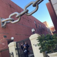 Urban art installation featuring a massive metallic chain draped across the top of a gated entrance between two white pillars. The chain connects adjacent buildings, suggesting themes of connection, tension, or unity. A person stands below with raised arms, gesturing toward the oversized chain. Decorative globe lamps perch atop the pillars, and a red-brick building flanks the scene under a clear sky. The Never Bust Chain