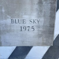 Concrete plaque mounted on a black-and-white diagonally striped wall. The plaque in Columbia, SC reads “BLUE SKY 1975” in engraved capital letters. A sliver of grass peeks from the bottom edge, adding a subtle touch of nature against the urban setting.