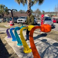 Person interacting with a colorful hand-played musical sculpture in Columbia, SC; featuring five curved chimes painted in vivid rainbow hues, arranged in a palm-lined park with cars and sculptures nearby.