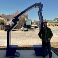 A striking blue metal sculpture shaped like a lightning bolt or jagged arch rises from a sunny urban sidewalk. A person stands beside it, enhancing the scale and interactive vibe. In the background, buildings, parked cars, and a gas station create a grounded, everyday setting, while the crisp blue sky adds visual contrast.